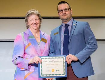 A woman in a pink and blue jacket and a man in a blue jacket and a checked shirt hold a certificate between them.