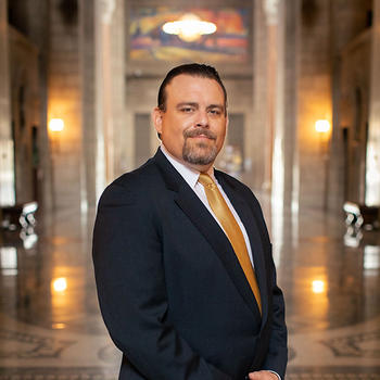 A distinguished looking gentleman with a dark goatee in a black suit and gold necktie stands in a regal room.